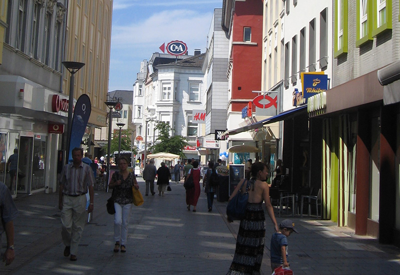 RME | Retail & Office | Timevest Portfolio – Menschen spazieren durch Einkaufsstraße in Innenstadt. Ladenzeilen mit Schaufenstern und Logos.